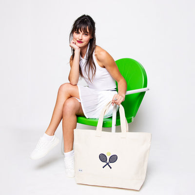 Woman sits on a green lawn chair on a white background. She it holding a country club tote canvas bag with crossed racquets embroidered on it