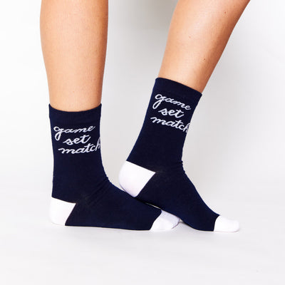 woman wears a pair of navy socks with white heel and toes. White text that reads game set match in cursive font around ankle