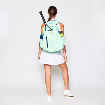 Woman stands on white background wearing white tennis dress and mint green tennis backpack.