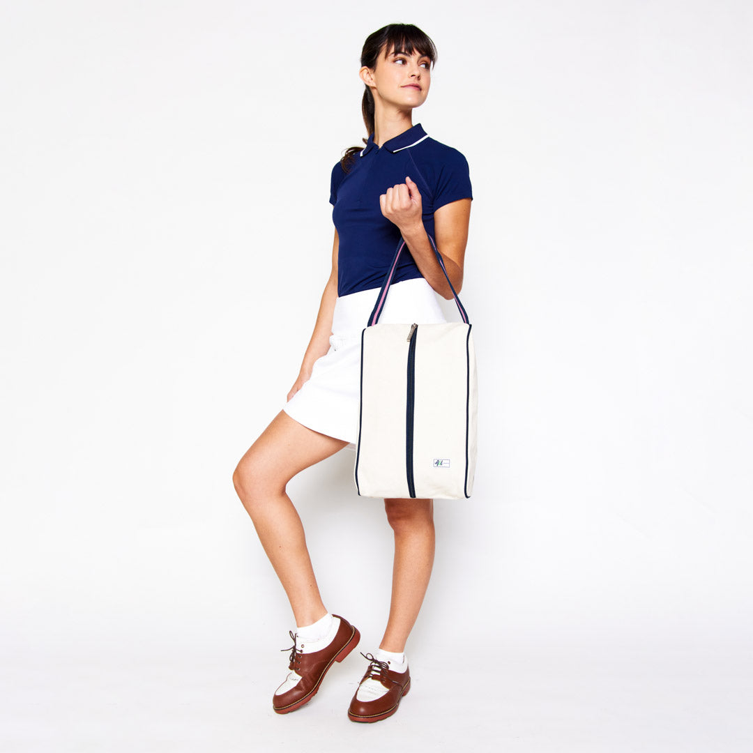 woman holds over her arm a rectangular tan canvas shoe bag with navy and pink handles.