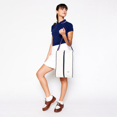 woman holds over her arm a rectangular tan canvas shoe bag with navy and pink handles.