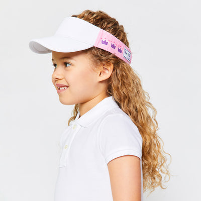 Little girl smiles wearing light pink kids visor with purple crowns on the side.