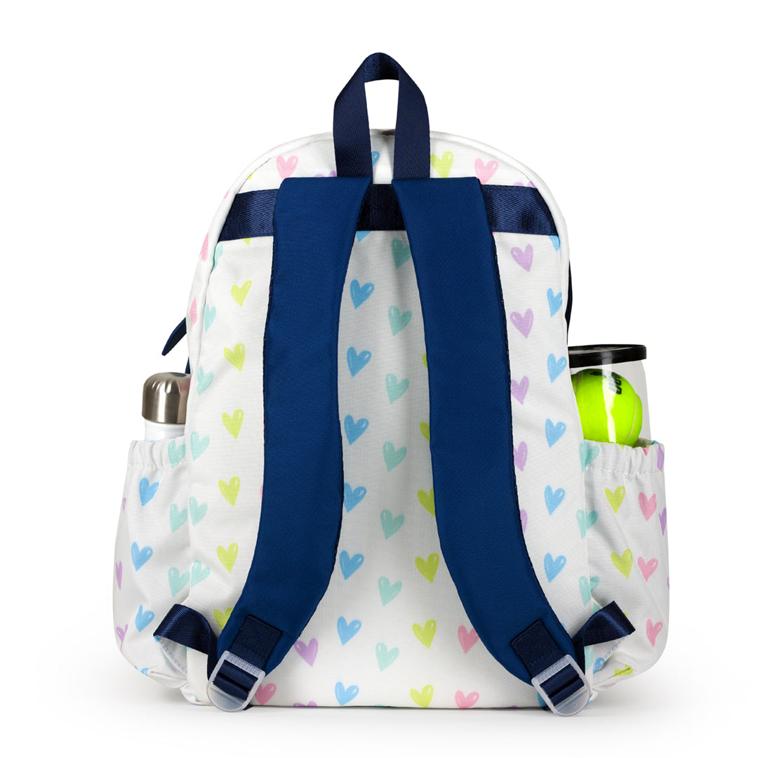 Back view of white kids tennis backpack with repeating pattern of rainbow hand drawn hearts. Bag has navy straps and trim