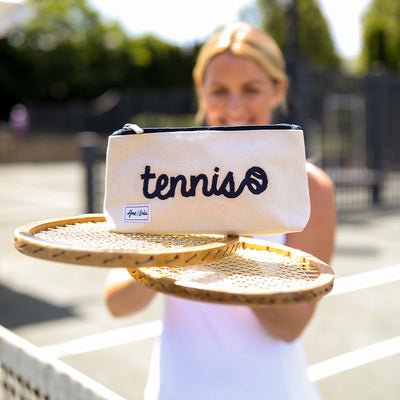 Woman stands on a tennis court with a racquet and makeup bag on top of racquet. small canvas makeup pouch with navy zipper. Front has the word tennis in cursive embroidered on.