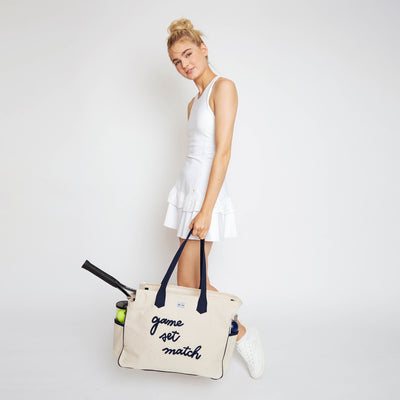 Woman stands on white background holding a love all court bag. The large canvas tennis tote reads "game set match" in a cursive font across the front of the bag.