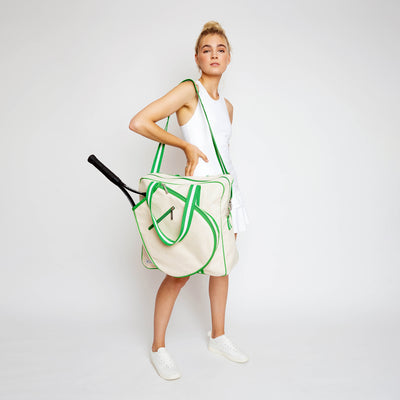 Woman stands on white background holding a canvas tennis tote in front of her. Tote has lime green trim and handles.