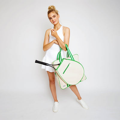 Woman stands on white background swinging a canvas tennis tote in front of her. Tote has lime green trim and handles.