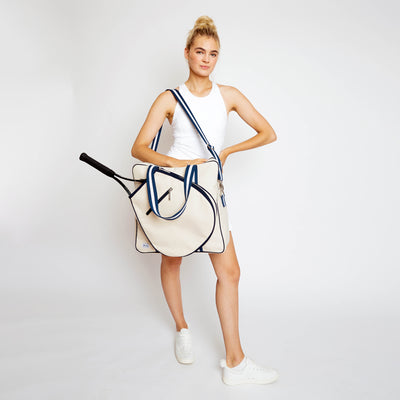 Woman stands on white background holding a canvas tennis tote in front of her. Tote has navy trim and handles.