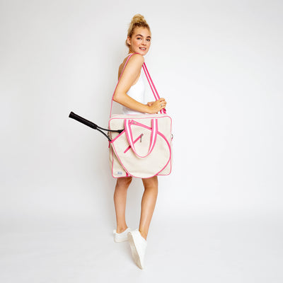 Woman stands on white background swinging a canvas tennis tote in front of her. Tote has hot pink trim and handles.