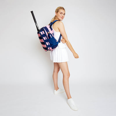 Woman stands on a white background with her side facing forward. She is wearing a navy and pink striped tennis backpack with a tennis racquet in the backpack front pocket.
