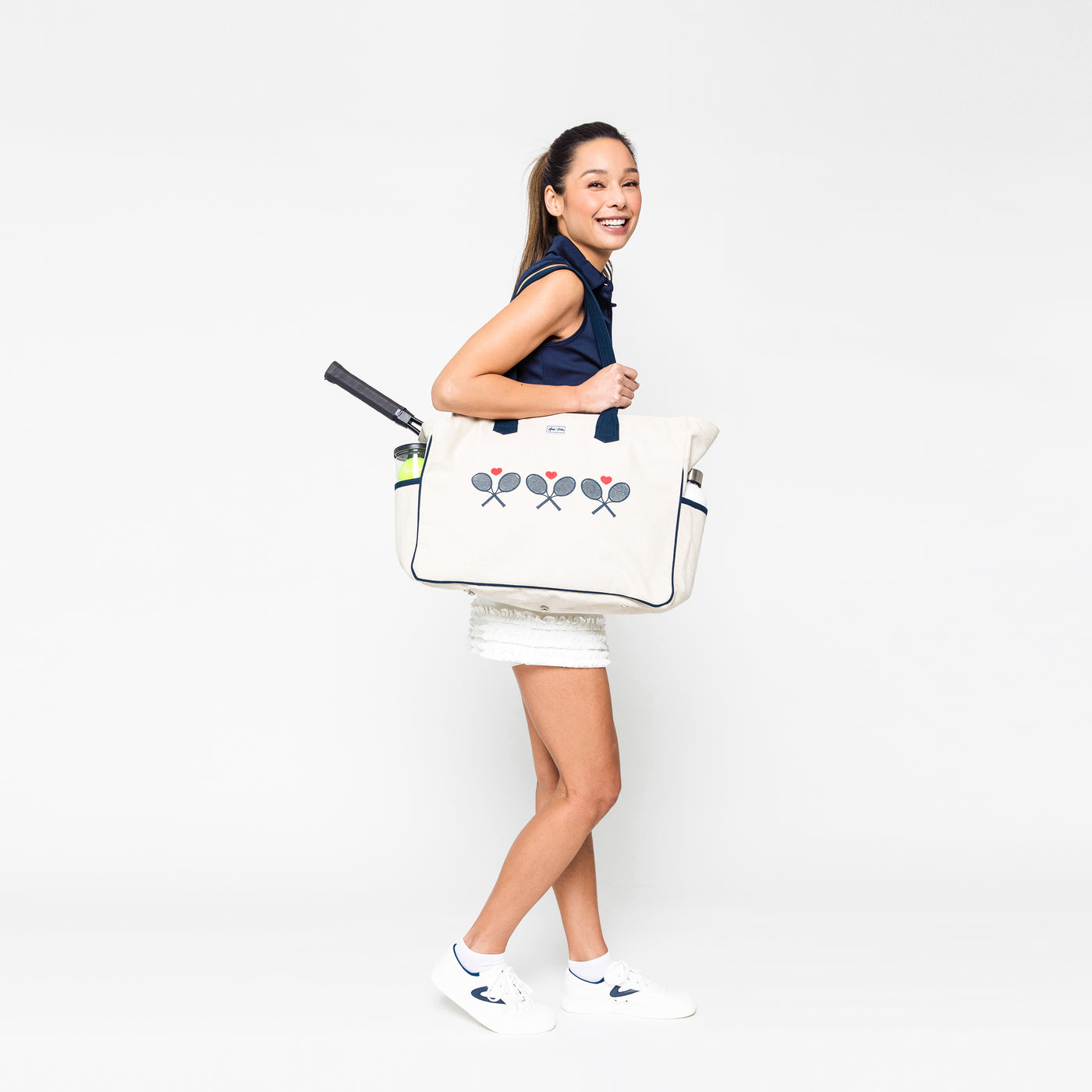 Woman stands on white background laughing and holding large canvas tennis tote. Tote has navy crossed racquets and red hearts printed across the front of it.
