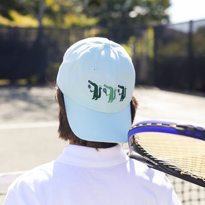 Boy on tennis court wearing light blue kids baseball hat with alligator playing tennis embroidered on the front backwards.