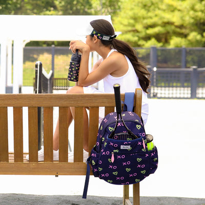 Girl sits on bench next to tennis court. navy kids tennis backpack with repeating green heart shaped tennis balls and pink x and o hangs on bench next to her.