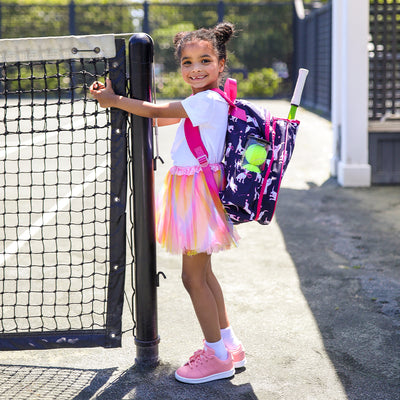 little girl stands by tennis net wearing navy kids tennis backpack with hot pink trim and pattern of white puppies holding tennis racquets and tennis balls