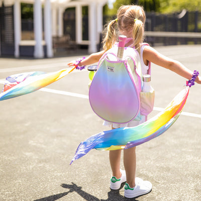 Little girl stands on tennis court waving rainbow scarves and wearing rainbow ombre kids tennis backpack.