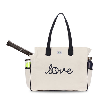 Front view of Love all court bag. Large canvas tennis tote with tennis racquet inside bag. Bag has side pockets holding water bottle and tennis balls. Front of bag reads the word love embroidered in a cursive font.