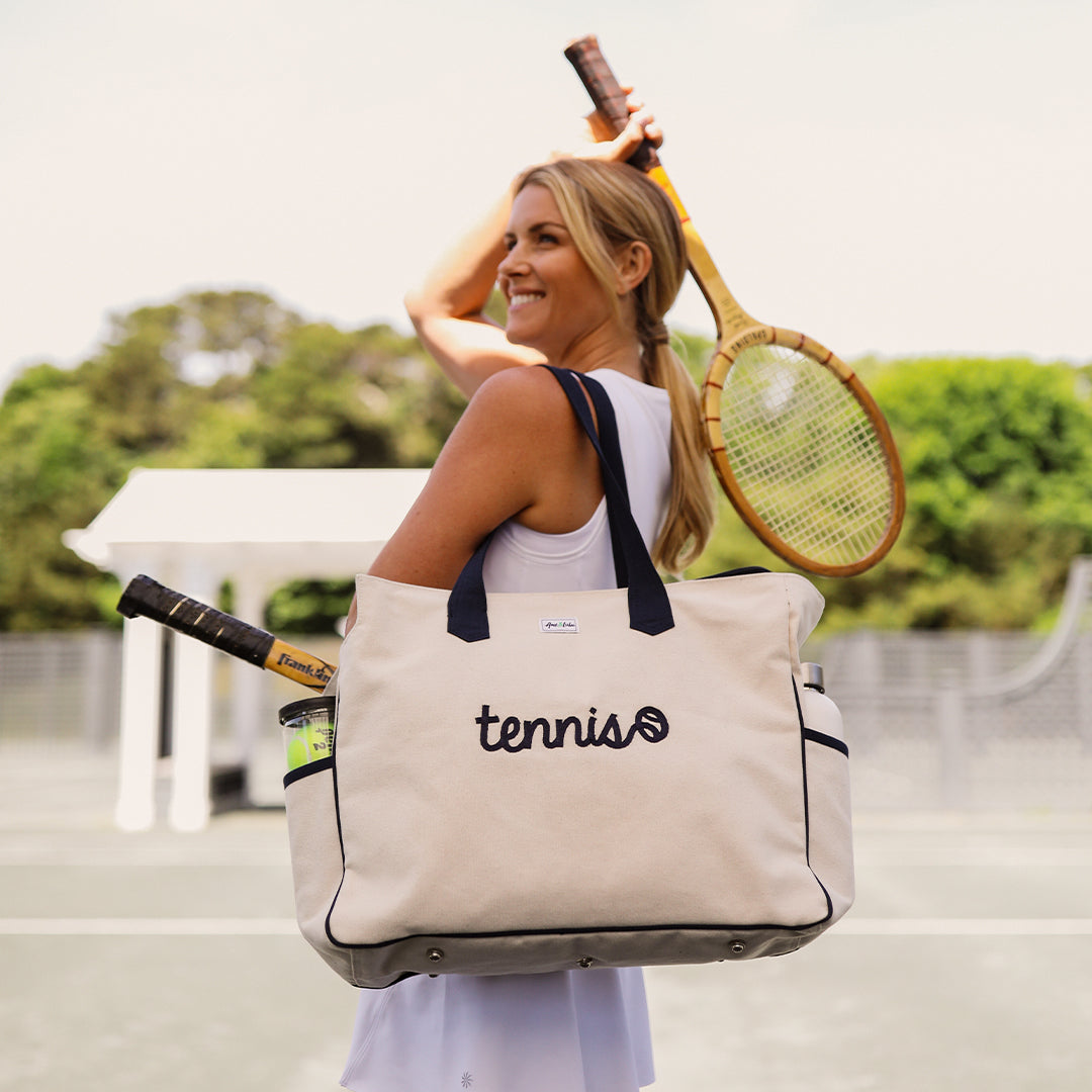 Woman stands on tennis court holding wooden tennis racquet and a love all court bag on her shoulder. Bag is a canvas bag that reads tennis in a cursive font on the front of it.