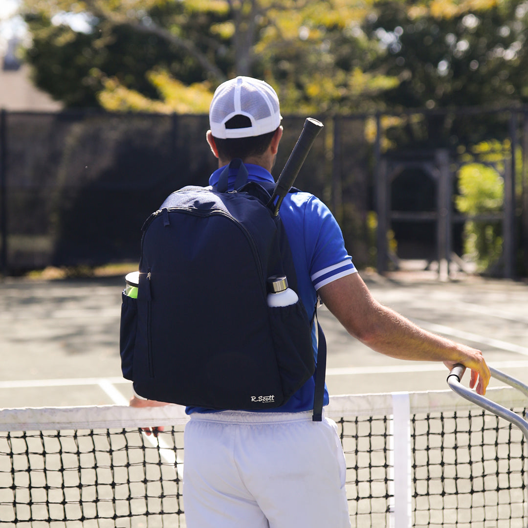 Man stands on tennis court next to tennis net wearing R Scott navy mens tennis backpack.