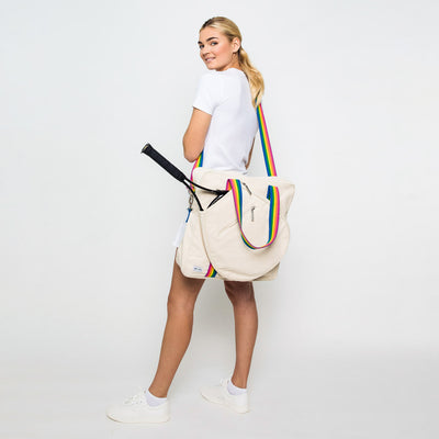 Woman stands on white background with canvas tennis tote over her shoulder. Tote has long rainbow strap.