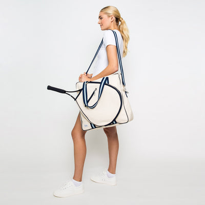 Woman stands on white background with hamptons tennis bag over her shoulder. Bag is large canvas tennis tote with navy and blue straps.
