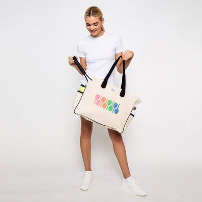 Woman stands on white background holding a canvas tennis tote with rainbow tennis racquets printed on front of bag.