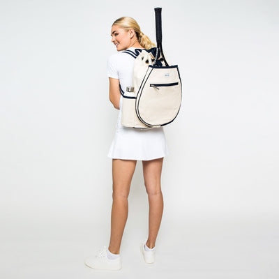 Woman stands on white background wearing canvas tennis backpack with navy trim.