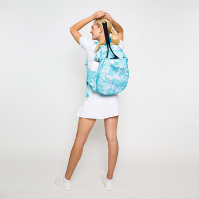 Woman stands on white background wearing aqua blue tie dye pattern tennis backpack.