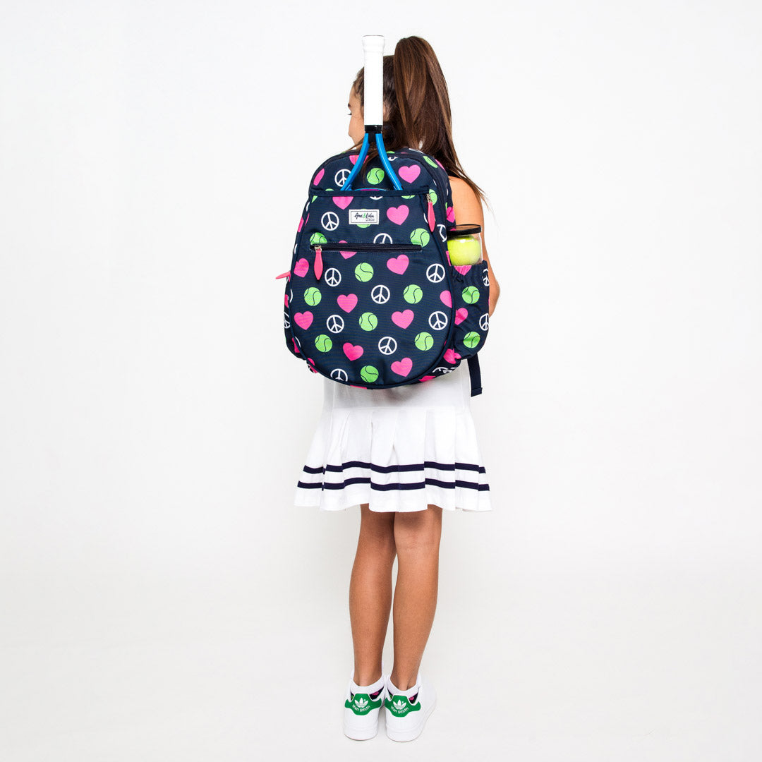 Girl stands on white background wearing navy kids tennis backpack with repeating pattern of white peace signs, pink hearts and green tennis balls.