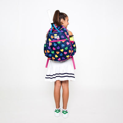 Little girl wearing navy kids tennis backpack with repeating rainbow heart shaped tennis balls pattern.