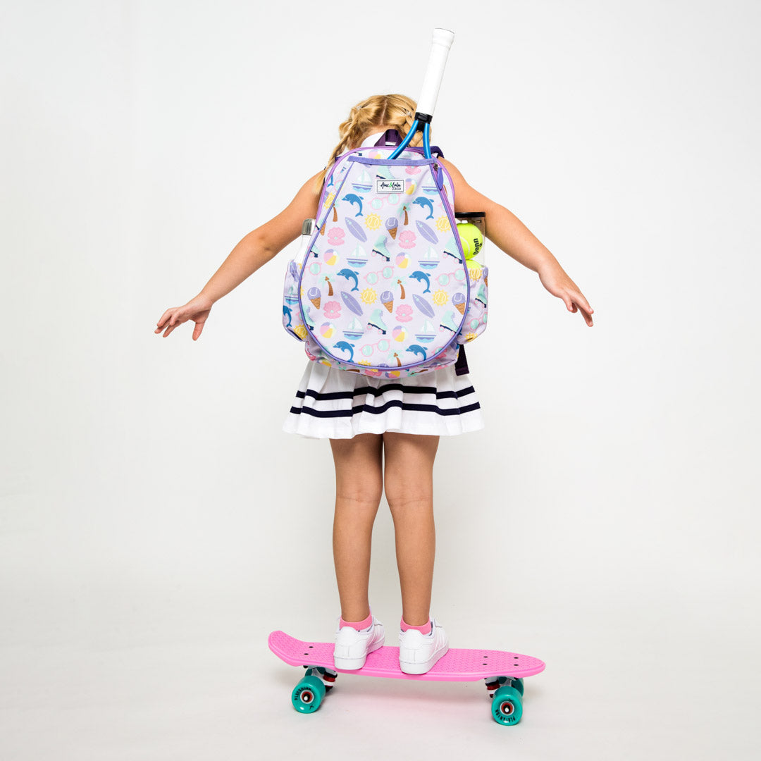 Little girl stands on skateboard wearing purple kids tennis backpack. Backpack is covered in beach themed icons such as surfboard, boats, beach ball and palm trees.