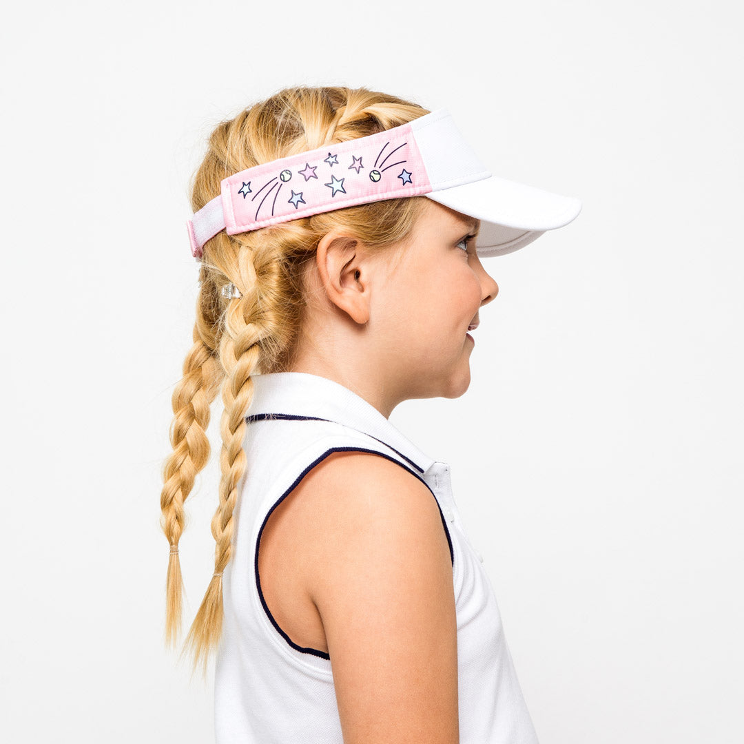 Little girl on white background wearing light pink kids visor with shooting stars and tennis balls printed on the sides.