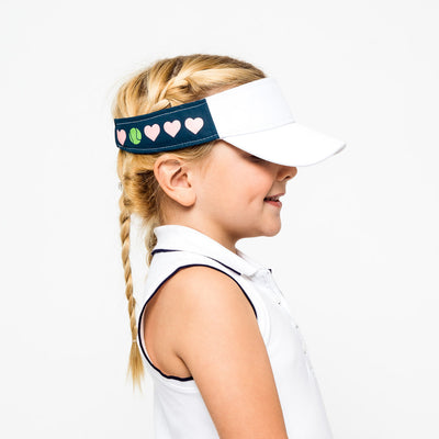 Little girl wearing navy kids visor with pink heart and green tennis balls printed on the sides.
