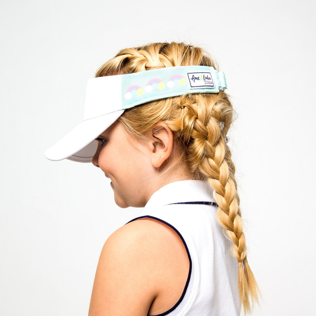 Little girl wearing light blue kids visor with rainbows and tennis balls printed on the sides.