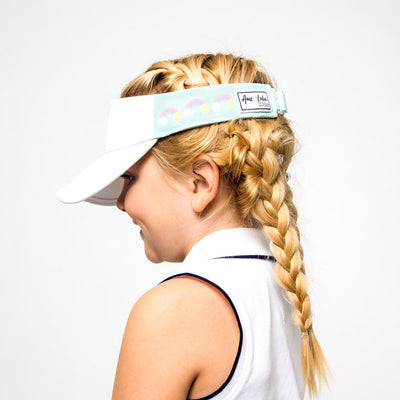 Little girl wearing light blue kids visor with rainbows and tennis balls printed on the sides.