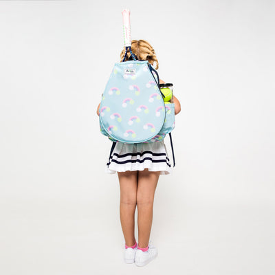 Little girl wearing a light blue kids tennis backpack with rainbow and tennis ball icons on the bag. There is a front pocket for holding tennis racquets.