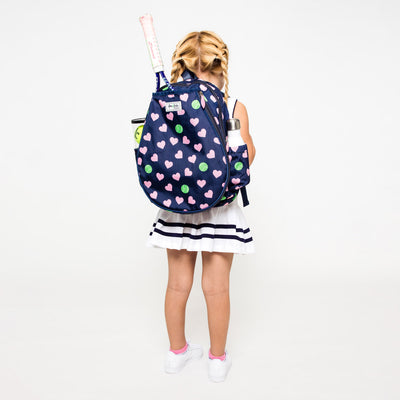 Little girl wearing a navy kids tennis backpack with pink hearts and green tennis ball pattern.