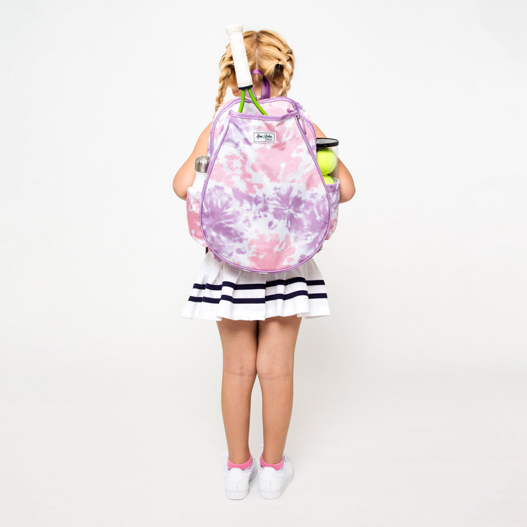 Little girl is wearing a pink and purple tie dye kids tennis backpack. There is a racquet pocket in the front holding a tennis racquet.