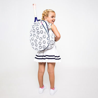 Little girl wearing a white kids tennis backpack with a navy tennis ball repeating pattern. Bag have navy trim and hot pink zippers.