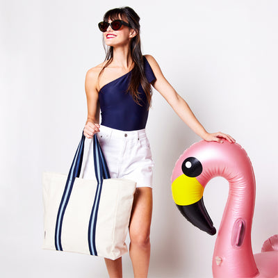 woman holding natural canvas tote with light blue and navy cotton webbing straps while leaning on a flamingo float