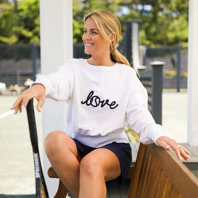 woman sits next to tennis court wearing white sweatshirt with the word love in cursive across front