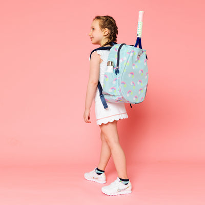 Little girl stands on a pink backdrop wearing a light blue kids tennis backpack with rainbow and tennis ball icons on the bag. There is a front pocket for holding tennis racquets.