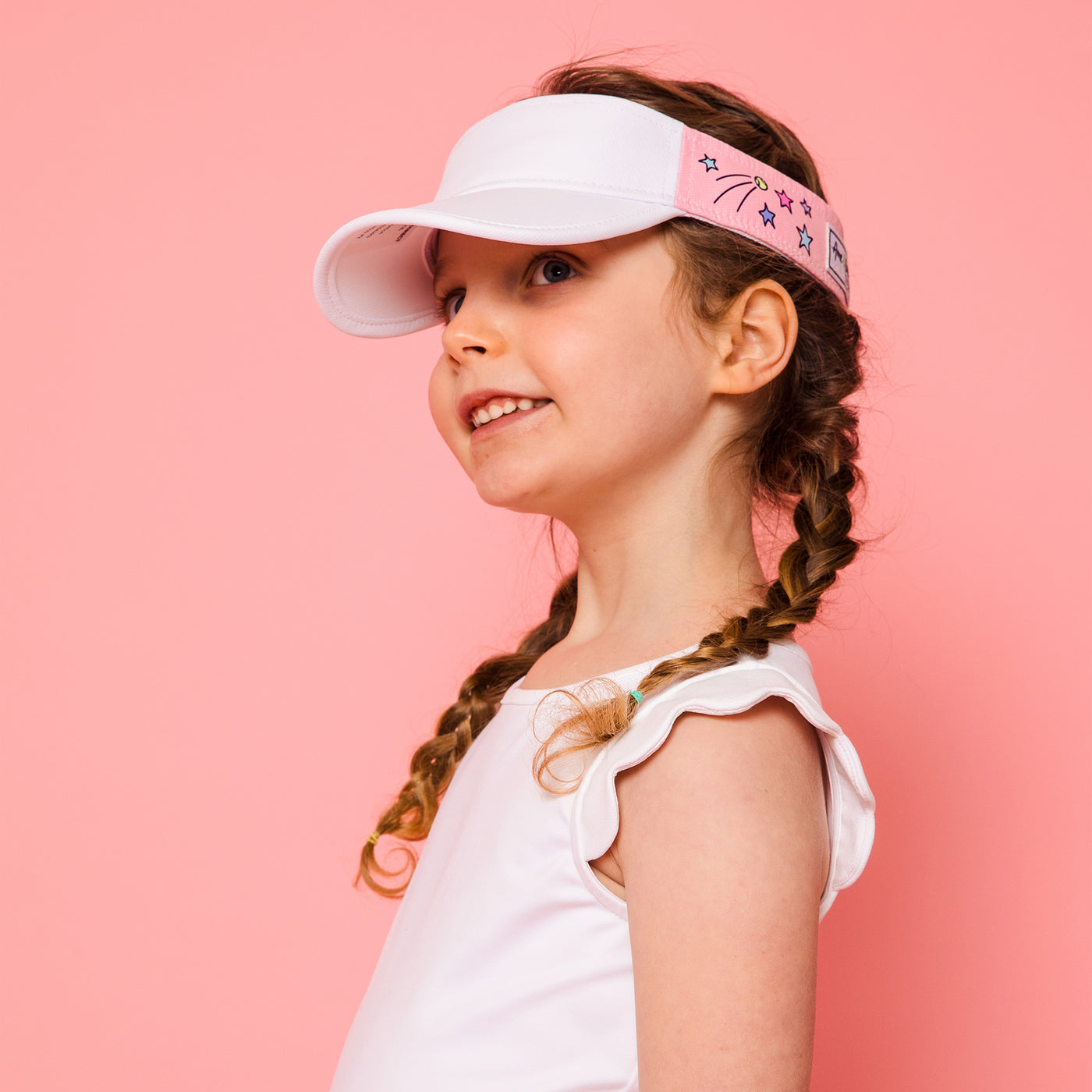 Little girl on pink background wearing light pink kids visor with shooting stars and tennis balls printed on the sides.