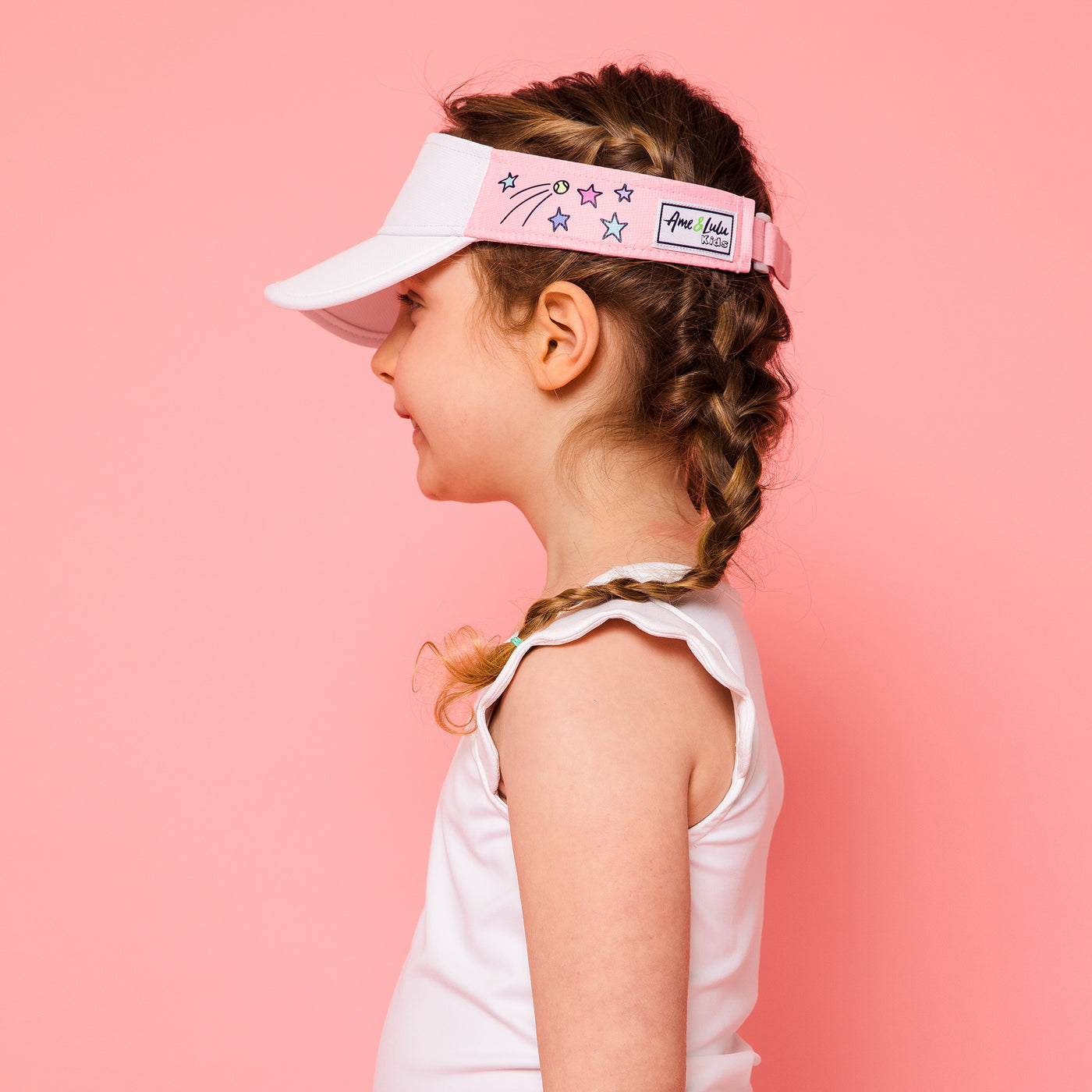 Little girl on pink background wearing light pink kids visor with shooting stars and tennis balls printed on the sides.