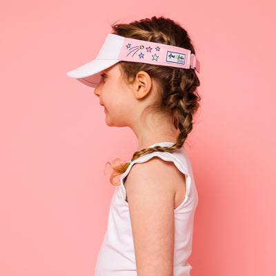 Little girl on pink background wearing light pink kids visor with shooting stars and tennis balls printed on the sides.
