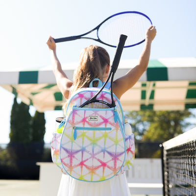 A girl stands on a tennis court holding a tennis racquet up in the air. She is wearing a white kids tennis backpack with rainbow shibori tie dye pattern.