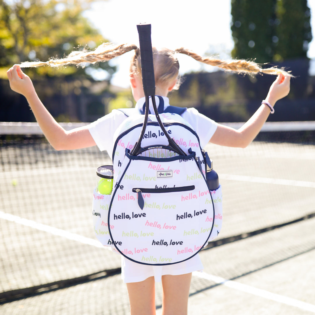Little girl standing on tennis court holding her braids above her head. She is wearing a white kids tennis backpack with repeating patterns of the words hello love in pink green and navy.