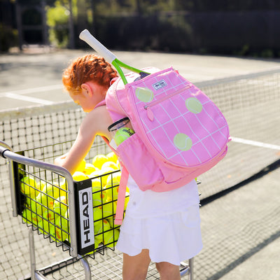 Little girl leans over tennis ball cart wearing a coral kids tennis backpack with repeating pattern of cream grid lines and yellow tennis balls.