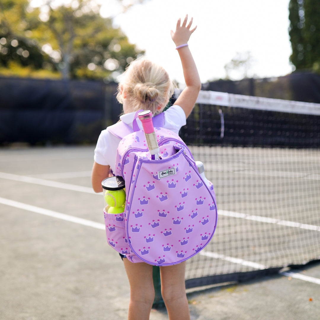 little girl stands on tennis court and wears a light pink kids tennis backpack with purple trim and purple crown repeating pattern