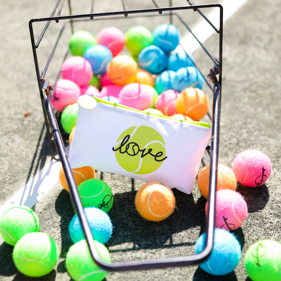 Basket of rainbow tennis balls tipped over on tennis court with white everyday pouch with lime green zipper is inside basket. Pouch has lime tennis ball with the word love in cursive printed on the front.