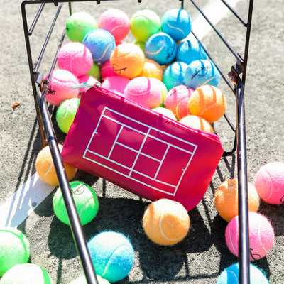 Basket of rainbow tennis balls is tipped over on a tennis court and a hot pink everyday pouch is in the basket. Pouch has white tennis court printed on front and lime green tennis racquet zipper pull.
